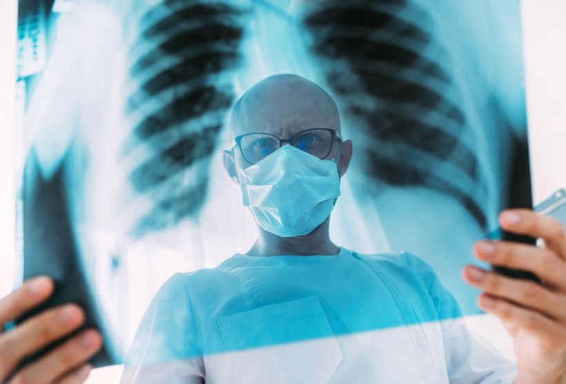 A doctor wearing glasses and a protective face mask is looking at the xray of a...